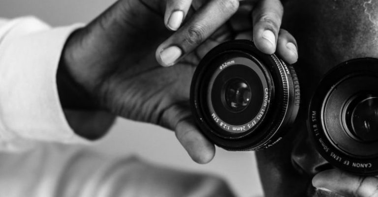 Digital Mystery - Black and white of faceless African American person covering eyes with lenses of photo camera on grey background