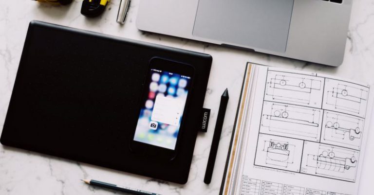 App Blueprint - From above view of modern workplace with laptop and tablet and smartphone placed near opened notebook with drawings and pen with pencil on white marble table