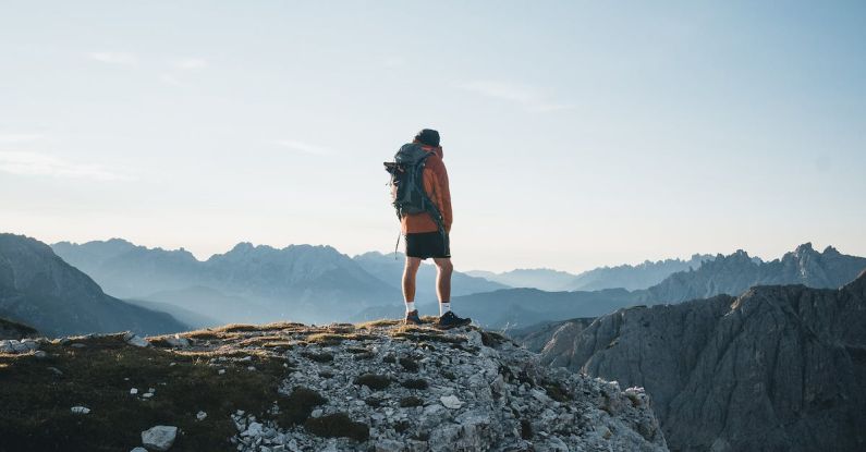 Climber Summit - Back view anonymous male explorer with backpack standing on rough mountain peak and enjoying breathtaking scenery of severe highland on clear day