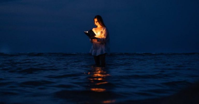 Books Horizon - A Woman Standing in the Water with a Book Illuminating Her Face